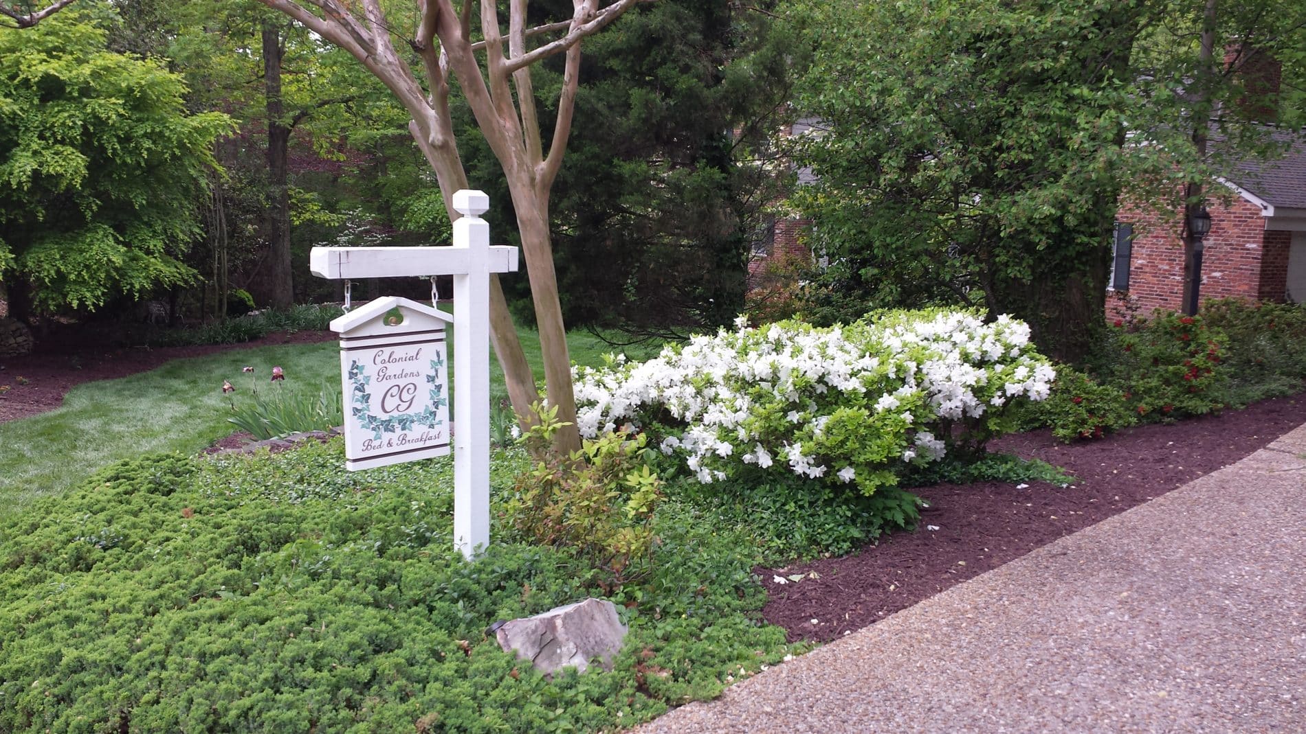 hanging wooden sign with the workds colonial gardens be and breakfast painted in white and green on yard with green lawn background