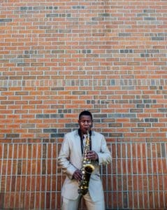 Man in light gray suit standing in front of a brick building playing a saxophone.