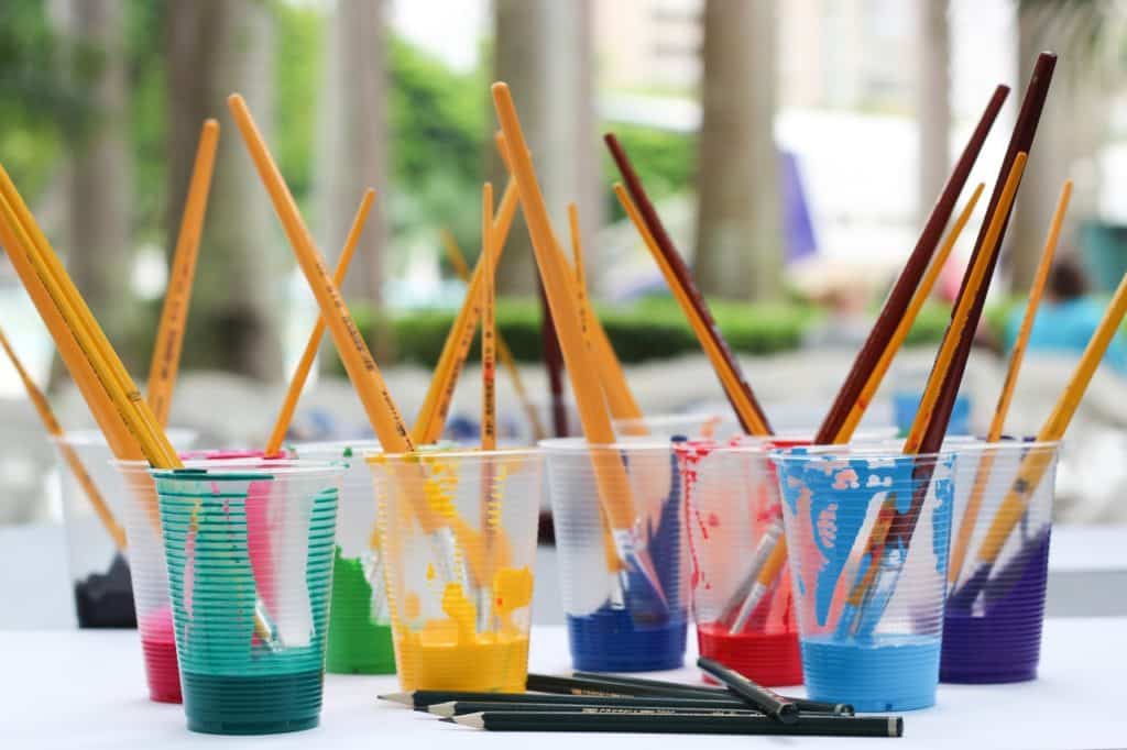 a table with multiple clear plastic cups each filled with a different colored paint and a paint brush