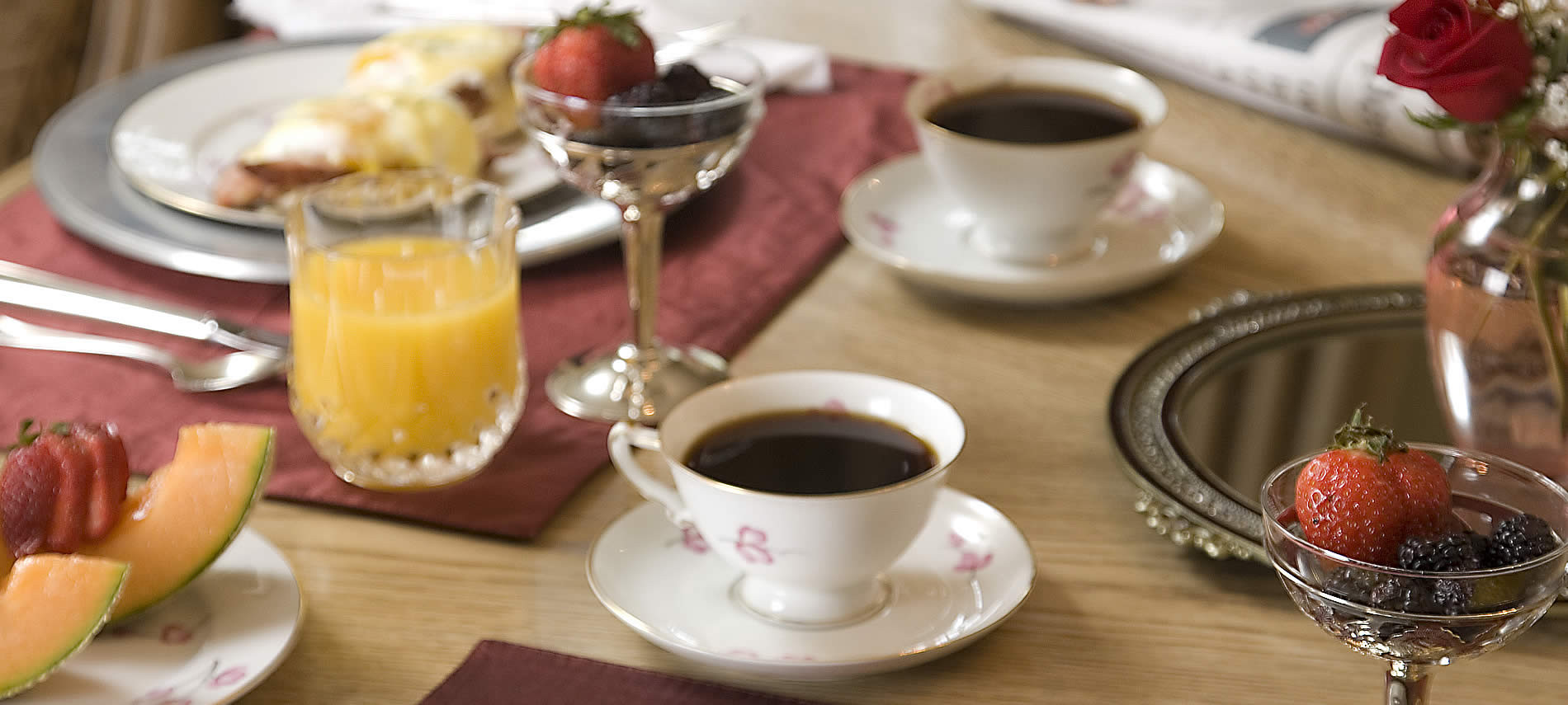 two coffee cups with saucer filled with coffee along side straberries in a glass and silver champagne glass on red placemat and glass of orange juice